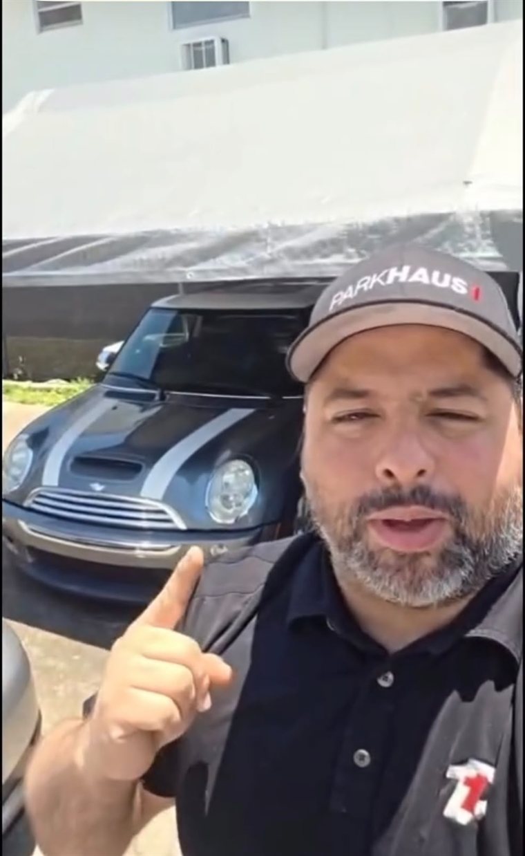Vinícius Lelis, ganhador da 1ª rifa, apontando para o MINI Cooper 2006 cinza com faixas prateadas que arrematou no leilão